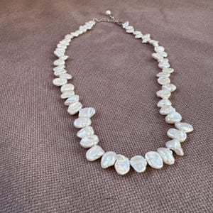 Puede incluir: Un collar de perlas blancas con perlas de forma irregular y orgánica. Las perlas están ensartadas en una cadena y un cierre de color plateado. El collar se muestra sobre una superficie marrón texturizada.