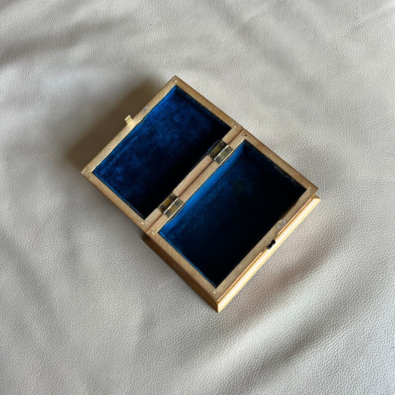 K&ouml;nnte beinhalten: Eine offene, rechteckige Holzbox mit messingfarbenem &Auml;u&szlig;eren und tiefblauem Samtinterieur. Die Box ist mit Scharnieren versehen und hat einen kleinen Verschluss. Die Box ist leer und liegt auf einer hellen Oberfl&auml;che.