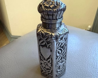 May include: A silver-colored perfume bottle with a floral design. The bottle has a rectangular shape with a decorative floral pattern and a heart-shaped emblem. The cap is also silver and has a flower-like design.