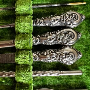Pode incluir: Um conjunto de ferramentas de manicure prateadas com al&ccedil;as ornamentadas, dispostas em uma caixa de veludo verde.