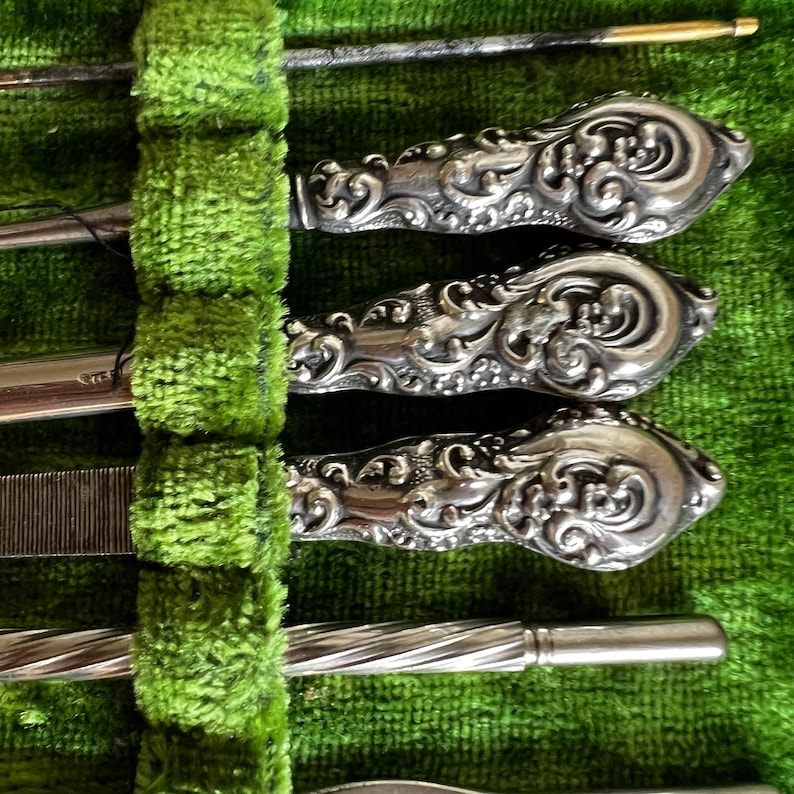 Pode incluir: Um conjunto de ferramentas de manicure prateadas com al&ccedil;as ornamentadas, dispostas em uma caixa de veludo verde.