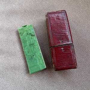 Op de afbeelding: Twee antieke rechthoekige dozen. Eén is een dieprode, scharnierende, lederen doos. De andere is een kleinere, open, groene doos met een stoffen bekleding met textuur. Beide dozen staan op een neutraal getinte, getextureerde ondergrond.