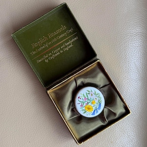 Puede incluir: Una caja verde vintage abierta con ribetes dorados y un compacto redondo de esmalte blanco. El compacto presenta un diseño floral pintado a mano con flores amarillas, azules y rosas. La caja dice "English Enamels" y "Enamelled on Copper and handpainted by Craftsmen in England."