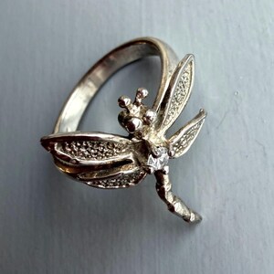 May include: Silver dragonfly ring with detailed wings and a small, clear gemstone. The ring features a detailed dragonfly design, with a polished finish. The ring is a piece of jewellery.