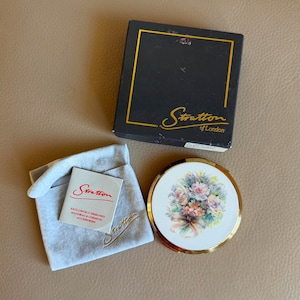 May include: A vintage Stratton of London compact with a floral design, gold trim, and a matching black box. The compact is accompanied by a grey pouch and a small card. The text on the box reads "Stratton of London".