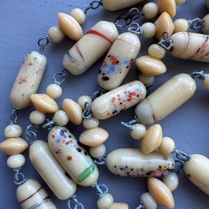 May include: A long necklace made of cream-coloured glass beads with colourful flecks and brown and white striped beads. The beads are strung on a thin cord with knots between each bead.