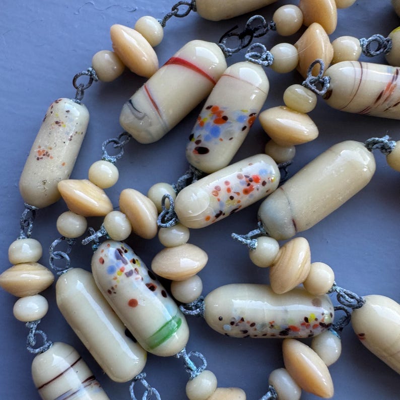 May include: A long necklace made of cream-coloured glass beads with colourful flecks and brown and white striped beads. The beads are strung on a thin cord with knots between each bead.