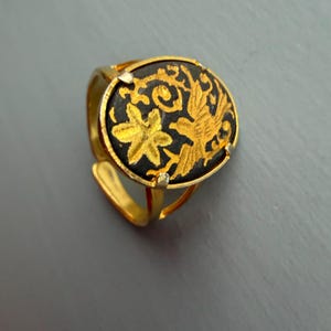 May include: Gold adjustable ring with a black circular face, adorned with a gold floral and bird motif. The ring has a plain band and a decorative setting around the central design.
