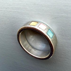 May include: A silver-toned band ring featuring a textured metallic finish and three small, square-shaped inlays in pale yellow, soft pink, and light blue, arranged in a row along the outer surface of the circular metal band.