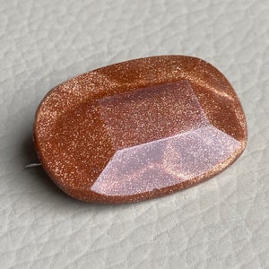 Puede incluir: Un broche ovalado de color marrón con un acabado brillante y brillante. El broche tiene un centro de vidrio transparente facetado.