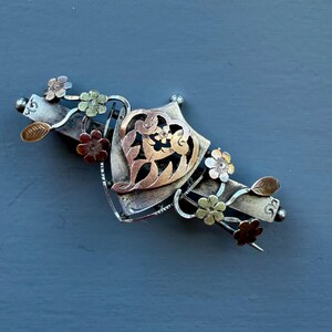 May include: A multi-toned metal brooch featuring a central shield-shaped element with an intricate heart-shaped cutout design. The piece is decorated with small, textured flowers and leaves in varying shades of silver, gold, and copper tones.