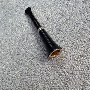 May include: A black smoking pipe with a silver band and a gold-colored rim. The pipe has a long, slender stem and a flared bowl. The pipe is made of a glossy material and is set against a textured gray background.