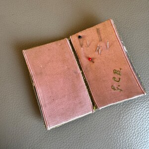 May include: A small, green and gold fabric sewing box with a pink interior. The box is open and has several pins inside. The box is embroidered with the initials "F.C.B."