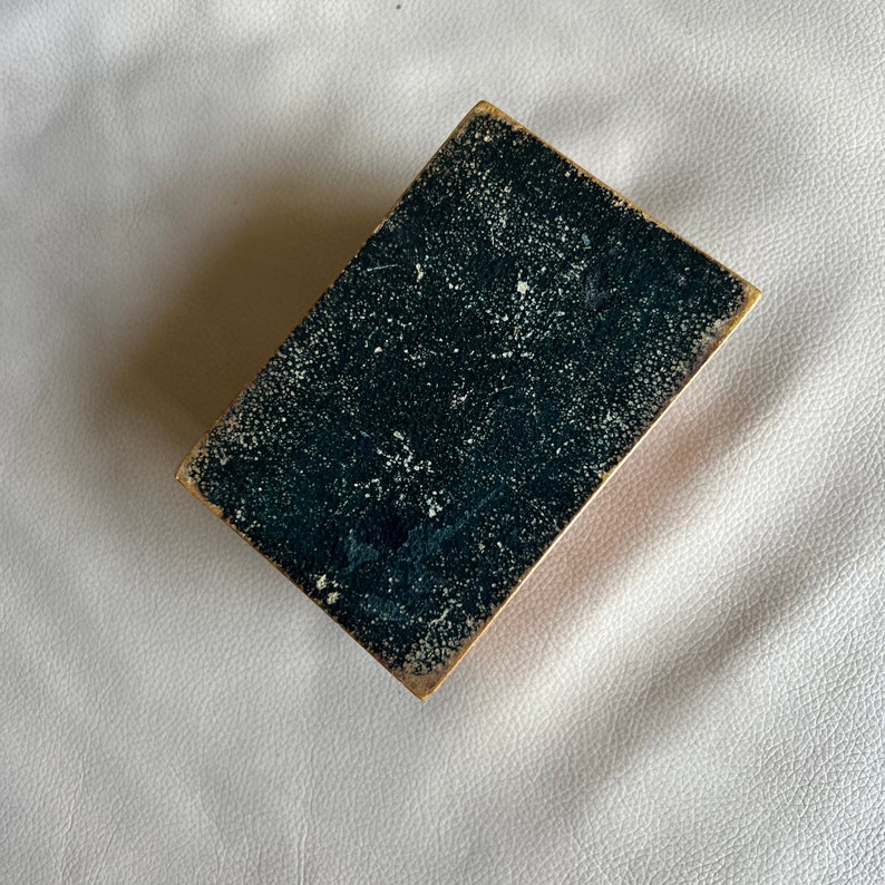 K&ouml;nnte beinhalten: Rechteckige Box im Vintage-Stil mit dunkelblau-gr&uuml;ner, gesprenkelter Oberseite und goldfarbenen Kanten. Die Box ist leicht angewinkelt und zeigt ihr gealtertes Aussehen und ihre Textur. Der Hintergrund ist hell und strukturiert.