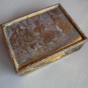 May include: A rectangular, gold-coloured box with an embossed scene of figures in period clothing. The lid features a detailed relief, and the box has ornate detailing around the edges. The box is closed with a small clasp.