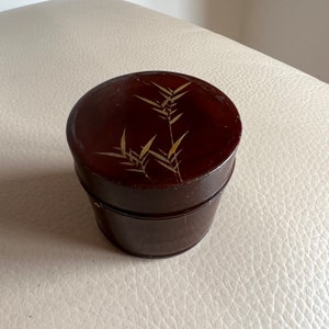 May include: A round, dark brown wooden box with a lid. The lid is decorated with a gold design of bamboo leaves.