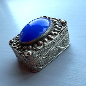 Puede incluir: Una caja rectangular plateada con una piedra ovalada azul brillante en la tapa. La caja está intrincadamente decorada con patrones florales y de cuentas. Probablemente para joyas o pequeños recuerdos.