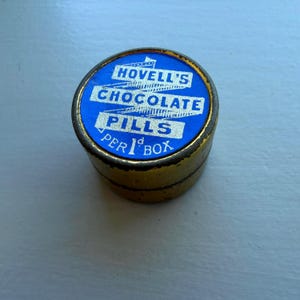 May include: A small, round, vintage tin with a blue and white label. The label reads "Hovell's Chocolate Pills" in white text. The tin is gold-colored with a slightly worn appearance, suggesting age.
