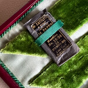 Pode incluir: Um pacote vintage de agulhas numa caixa de veludo verde. O pacote est&aacute; etiquetado "W. Bartleet & Sons Calforsted 1d. Paper Needles No. 18/24 -12- Needles London Made in England."