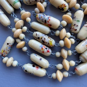 May include: A multi-strand necklace made of cream-coloured glass beads with colourful flecks and cream-coloured wooden beads. The beads are strung on a grey cord.