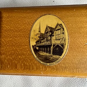 K&ouml;nnte beinhalten: Rechteckige Holzbox mit strukturierter Oberfl&auml;che. Ein ovales Bild auf dem Deckel zeigt eine schwarz-wei&szlig;e Illustration eines Geb&auml;udes mit Turm und dem Text "MARKET HOUSE MUCH WENLOCK". Die Box hat eine hellbraune Farbe.