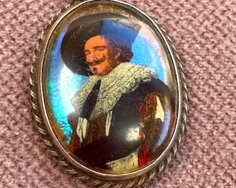 May include: An oval pendant featuring a portrait of a person wearing a wide-brimmed hat and a ruffled collar, set within a twisted silver-tone metal frame. The background of the portrait displays iridescent blue tones characteristic of butterfly wing.