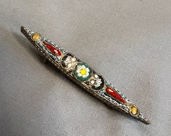 May include: A silver brooch with a long, narrow design. The brooch is decorated with colorful mosaic tiles in shades of red, green, white, and black. The center of the brooch features a white flower with a yellow center.