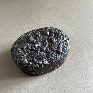 May include: An oval-shaped, dark brown trinket box with an intricate floral design embossed on the lid.