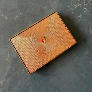 May include: A rectangular gold-tone metal compact case featuring a textured engine-turned surface with a geometric pattern. The center of the lid displays a small, detailed floral motif of a single pink rose with green leaves on a gold background.