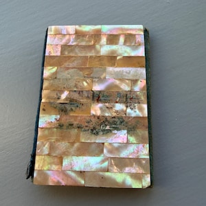 Puede incluir: Un pequeño cuaderno rectangular con una cubierta hecha de baldosas rectangulares iridiscentes de nácar. Las baldosas están dispuestas en un patrón de ladrillo. El lomo es de tela azul oscuro. El cuaderno está cerrado.