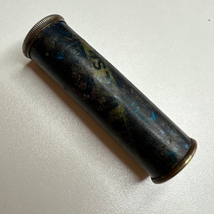 Puede incluir: Un tubo de metal negro y dorado con una etiqueta que dice "First Aid" en letras azules. El tubo tiene un aspecto desgastado y envejecido.