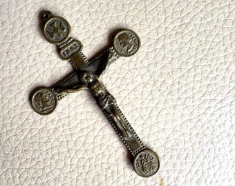 May include: A bronze-toned metal crucifix pendant featuring a central figure of Jesus on the cross. The cross arms terminate in circular medallions depicting religious figures and symbols, with a small loop at the top for attachment to a chain.