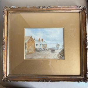 May include: A framed watercolour painting of a rural scene, featuring buildings, a road, and a horse-drawn cart. The painting is set within a gold-toned frame with decorative detailing, presenting a vintage aesthetic.