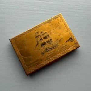 May include: A rectangular, gold-colored compact with a detailed engraving of a sailing ship on the lid. The ship is depicted with multiple sails and is set against a backdrop of stylized clouds and water. The edges of the compact have a textured border.