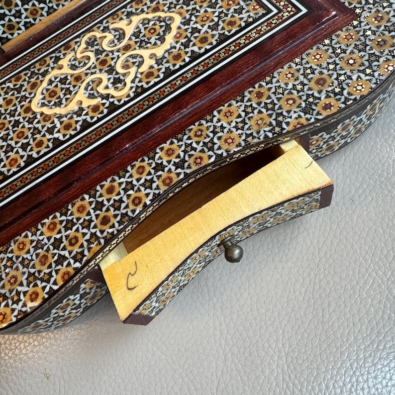 Pode incluir: Um close-up detalhado de uma caixa de madeira com uma gaveta aberta. A caixa apresenta intrincados padr&otilde;es de mosaico em marrom, dourado e branco. A gaveta &eacute; feita de madeira clara com um pequeno bot&atilde;o de metal. A caixa &eacute; provavelmente para joias ou itens pequenos.