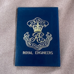 May include: A dark blue booklet featuring gold embossed text and a crest. The crest includes a crown, the letters "RE", and the words "Ubique" and "Royal Engineers". The booklet is rectangular and closed.