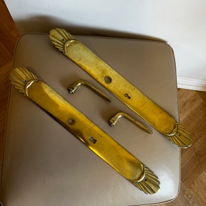 May include: Two brass door handles with decorative scalloped edges and two matching brass door knobs. The handles are long and rectangular with a keyhole and a round hole for a screw. The knobs are simple and curved.