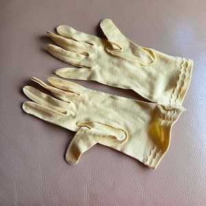 May include: A pair of pale yellow gloves with a scalloped edge. The gloves are made of a soft fabric and have a classic design. The gloves are laid out on a neutral surface.