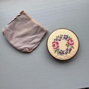 May include: A vintage compact with a floral cross-stitch design in pink, blue, and purple. The compact is round with a gold-tone metal frame. A matching lavender pouch is next to the compact.