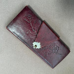 Puede incluir: Una cartera rectangular de cuero color burdeos oscuro con cierre de solapa diagonal y un broche de metal plateado. La superficie exterior presenta una textura entrecruzada y está grabada con un patrón floral de tallos y flores.