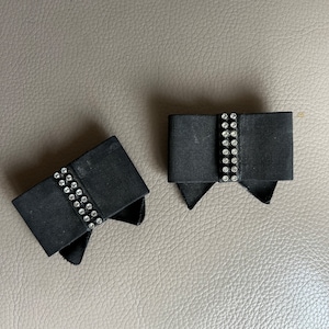 May include: Two black fabric bow clips with rows of clear rhinestones. The bows are tied in a simple knot and have pointed ends.