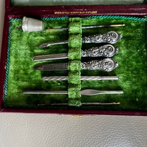 Pode incluir: Uma caixa vintage forrada de veludo verde com uma inscri&ccedil;&atilde;o prateada "WARRANTED SHEFFIELD CUTLERY" na tampa. A caixa cont&eacute;m um conjunto de ferramentas de manicure com cabo prateado, incluindo uma lima de unha, uma pin&ccedil;a e um empurrador de cut&iacute;culas.
