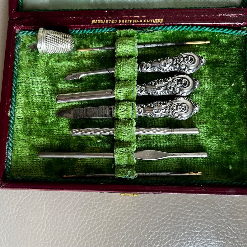 Pode incluir: Uma caixa vintage forrada de veludo verde com uma inscri&ccedil;&atilde;o prateada "WARRANTED SHEFFIELD CUTLERY" na tampa. A caixa cont&eacute;m um conjunto de ferramentas de manicure com cabo prateado, incluindo uma lima de unha, uma pin&ccedil;a e um empurrador de cut&iacute;culas.