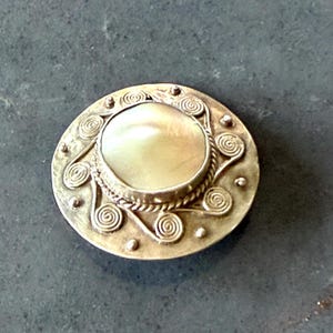 May include: A round brooch featuring a central iridescent mother-of-pearl cabochon set in a metallic frame. The border is decorated with a series of raised metal spirals and small spherical studs arranged in a circular pattern around the center.