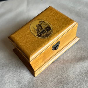 K&ouml;nnte beinhalten: Eine rechteckige Holzkiste mit hellbraunem Finish. Die Kiste hat ein dekoratives ovales Bild auf dem Deckel und einen Metallverschluss. Das Bild auf dem Deckel zeigt ein Geb&auml;ude mit dem Text "Market House, Much Wenlock".