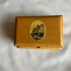 K&ouml;nnte beinhalten: Eine rechteckige Holzkiste mit hellbraunem Finish. Der Deckel zeigt ein ovales Bild eines Geb&auml;udes mit dem Text "MARKET HOUSE MUCH WENLOCK". Die Kiste ist geschlossen.