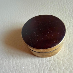 May include: A small, round, lidded container featuring a dark brown, polished wooden top surface and a metallic, gold-toned base. The cylindrical object has a smooth, flat lid that sits flush with the sides of the base, creating a simple, compact form.