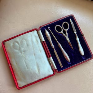 Könnte beinhalten: Ein Vintage-Maniküre-Set in einem roten Etui mit blauem Samtinterieur. Das Set enthält eine kleine Schere, eine Nagelfeile, einen Nagelhautschieber und einen Häkelhaken, alle mit aufwendigen Silberdetails. Das Etui ist geöffnet und zeigt die Werkzeuge.