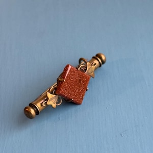 Puede incluir: Un broche dorado con una piedra cuadrada de color marrón rojizo y detalles decorativos.