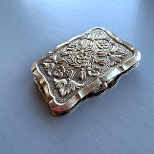 May include: An antique silver-coloured rectangular box with a floral design. The box has a detailed embossed pattern of roses, leaves, and other floral elements. The edges are slightly rounded, and the box appears to be closed.